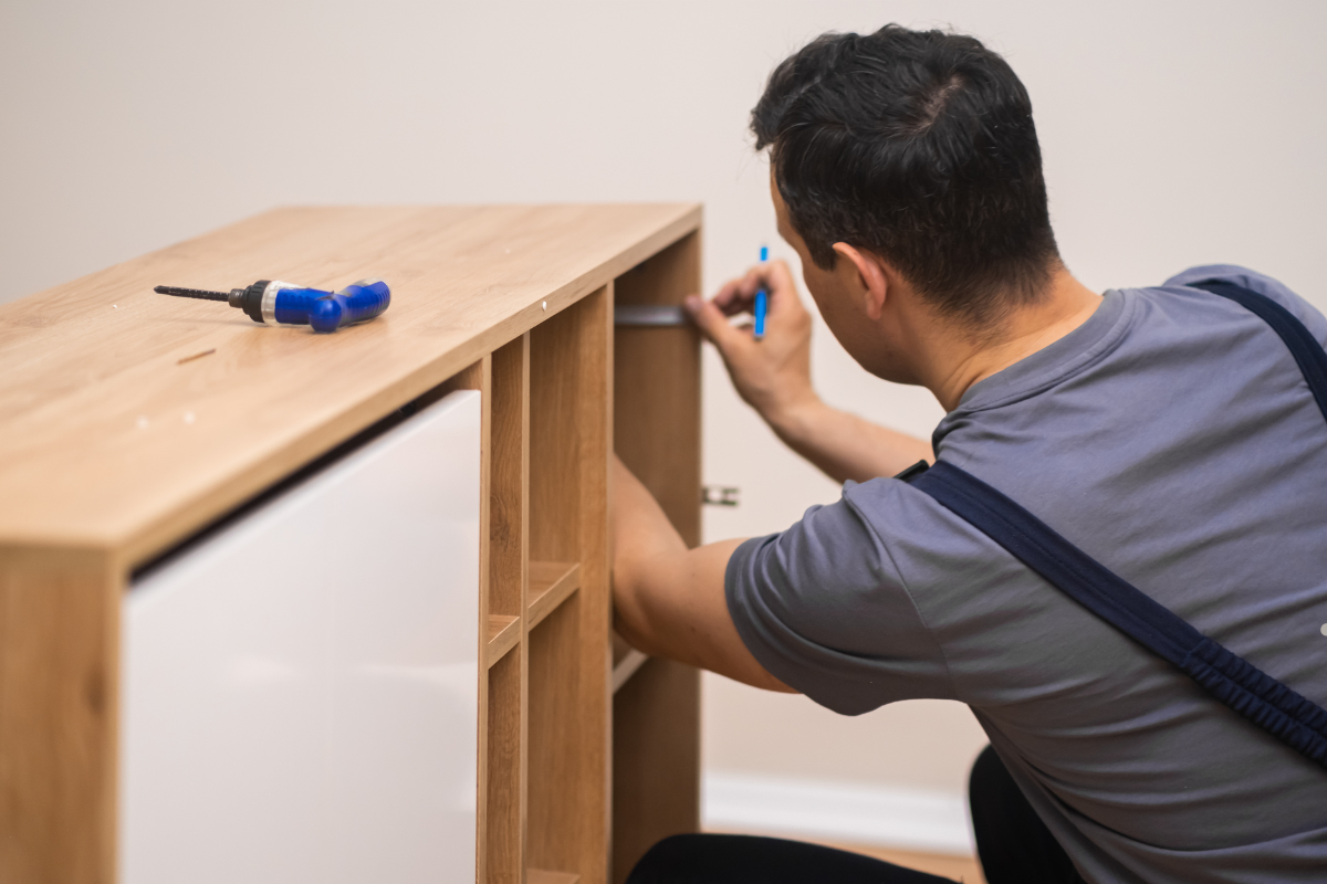 A person assembles a wooden shelf with visible compartments, using a screwdriver, on a smooth floor against a light-colored wall.