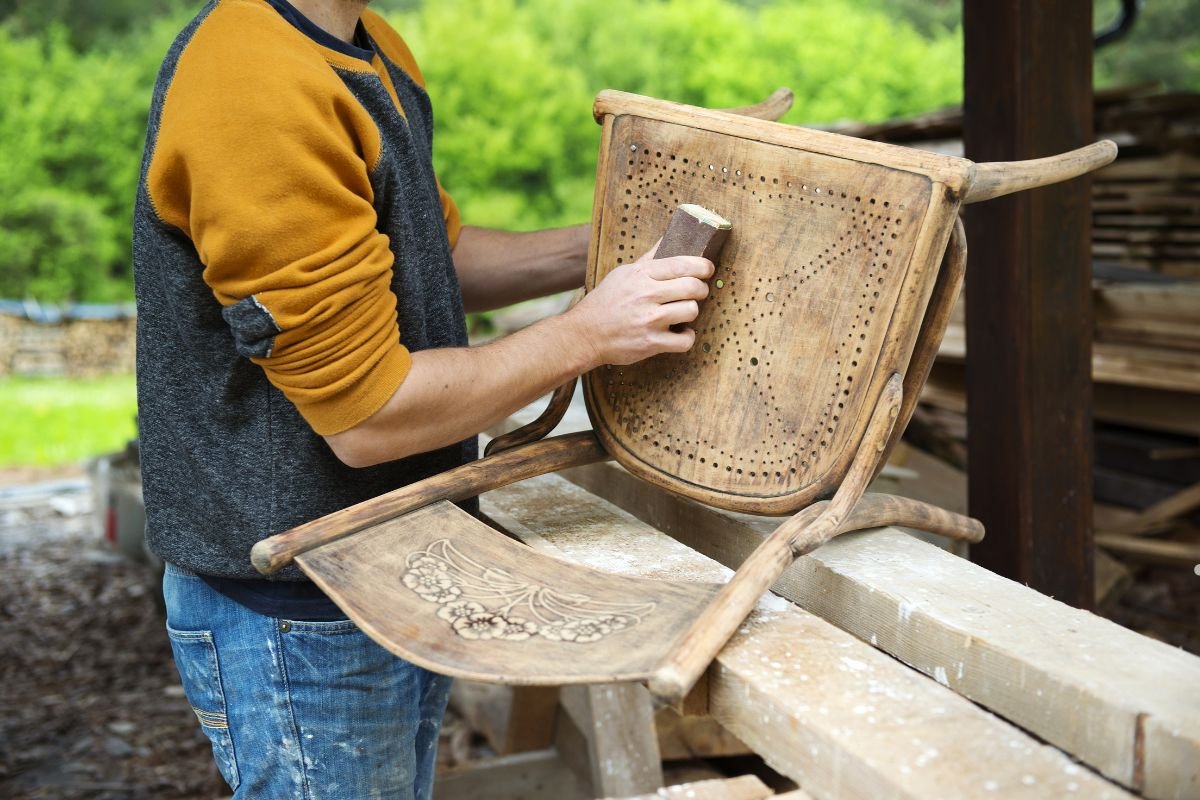 A person sands a wooden chair in a workshop, focusing on the intricately carved seat, surrounded by greenery and wood piles.