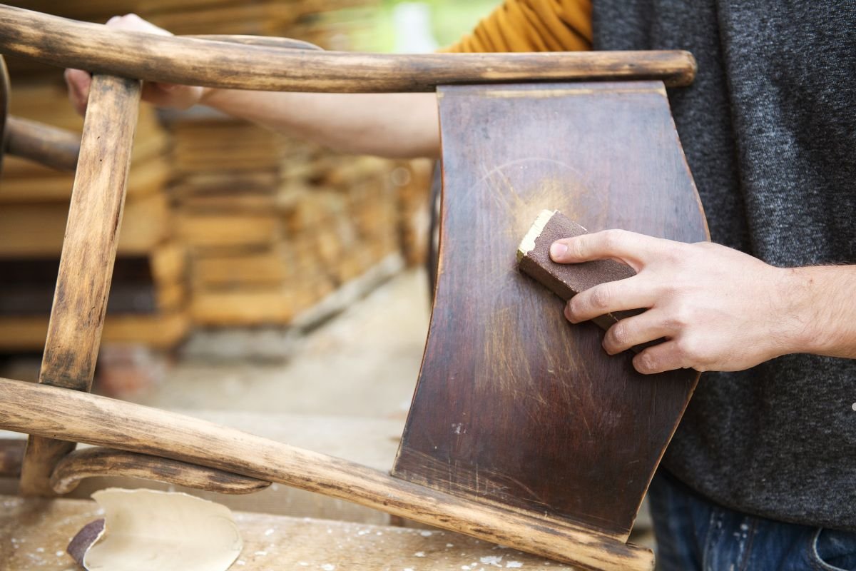 A person sands a rustic wooden chair, focusing on a curved backrest, surrounded by stacks of lumber in a workshop setting.