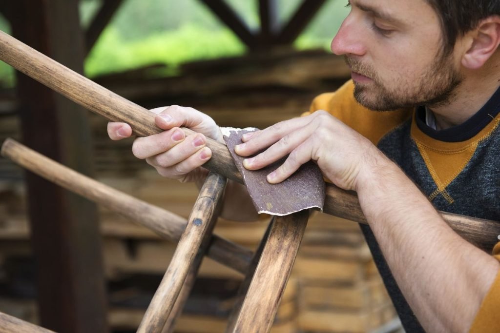 A person's hands hold a piece of sandpaper, smoothing a wooden structure under a shaded outdoor area.