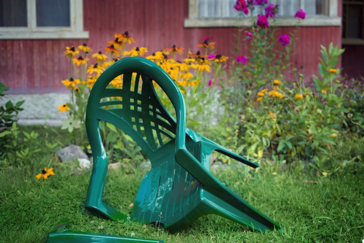 A broken chair lies on the grass, its wooden frame splintered and one leg missing, surrounded by green blades of grass.
